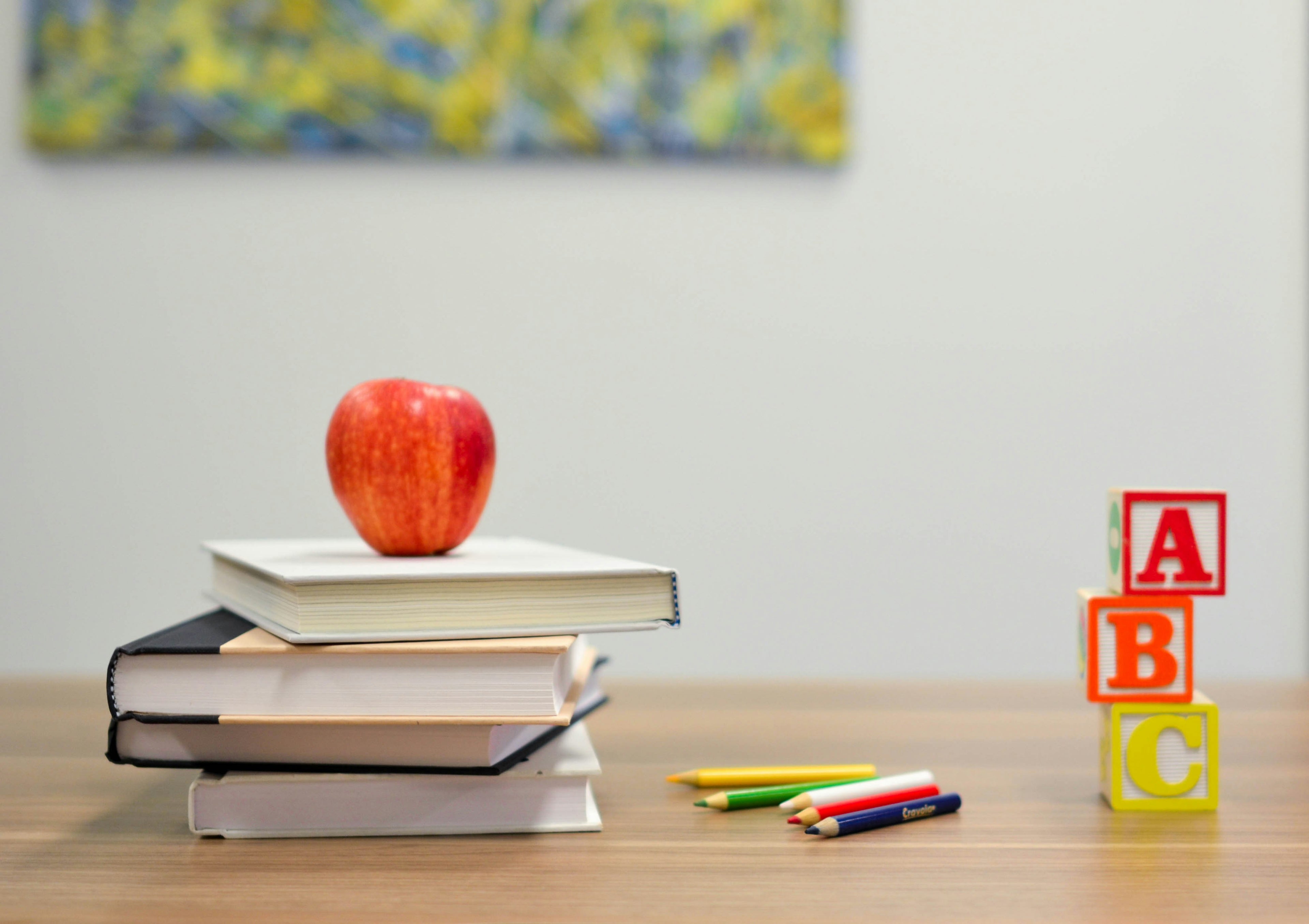Stack of books with an apple on top, surrounded by colorful pencils and ABC blocks on a wooden surface.